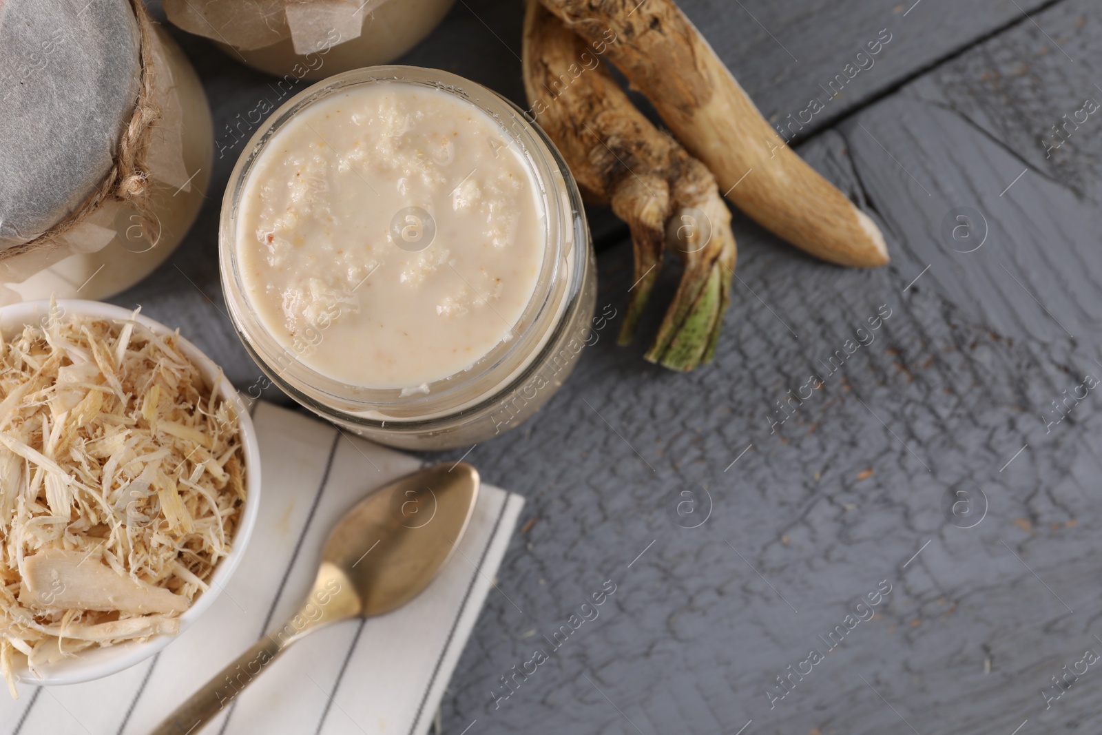Photo of Spicy horseradish sauce in jars, roots and spoon on grey wooden table, flat lay. Space for text
