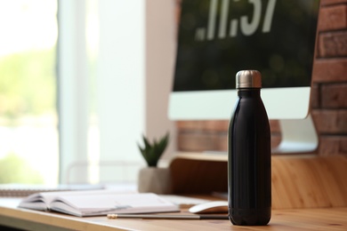 Photo of Modern black thermos bottle on wooden desk at workplace. Space for text