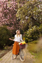 Beautiful young woman with bicycle and flowers in park on pleasant spring day