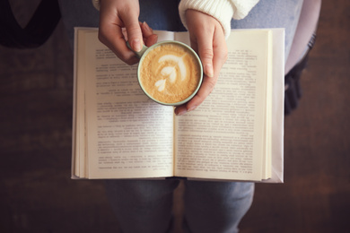 Photo of Woman with cup of coffee reading book indoors, top view