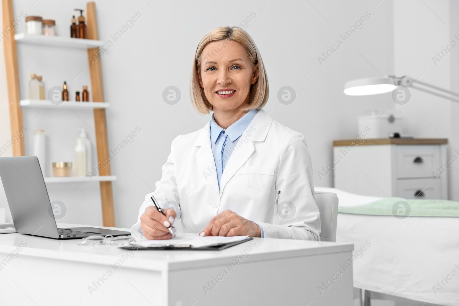 Photo of Portrait of happy dermatologist with pen at white table in clinic