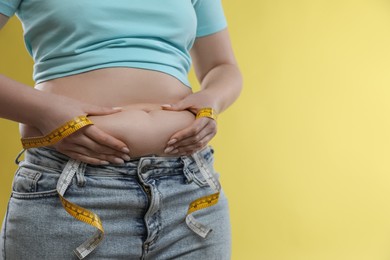 Woman with measuring tape touching belly fat on yellow background, closeup. Overweight problem