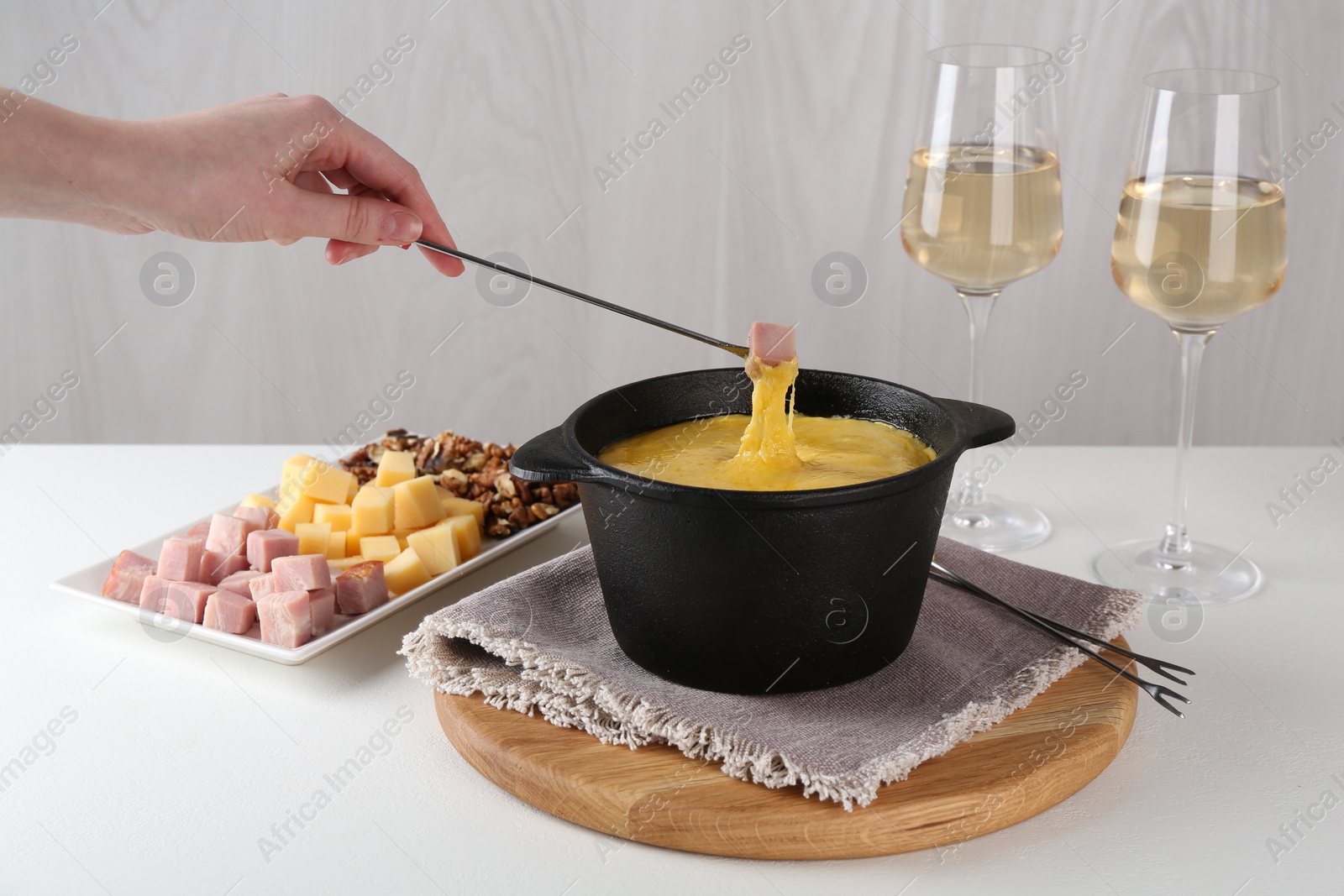 Photo of Dipping piece of ham into fondue pot with tasty melted cheese at white table, closeup