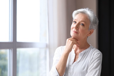 Happy mature woman near window indoors, space for text. Smart aging