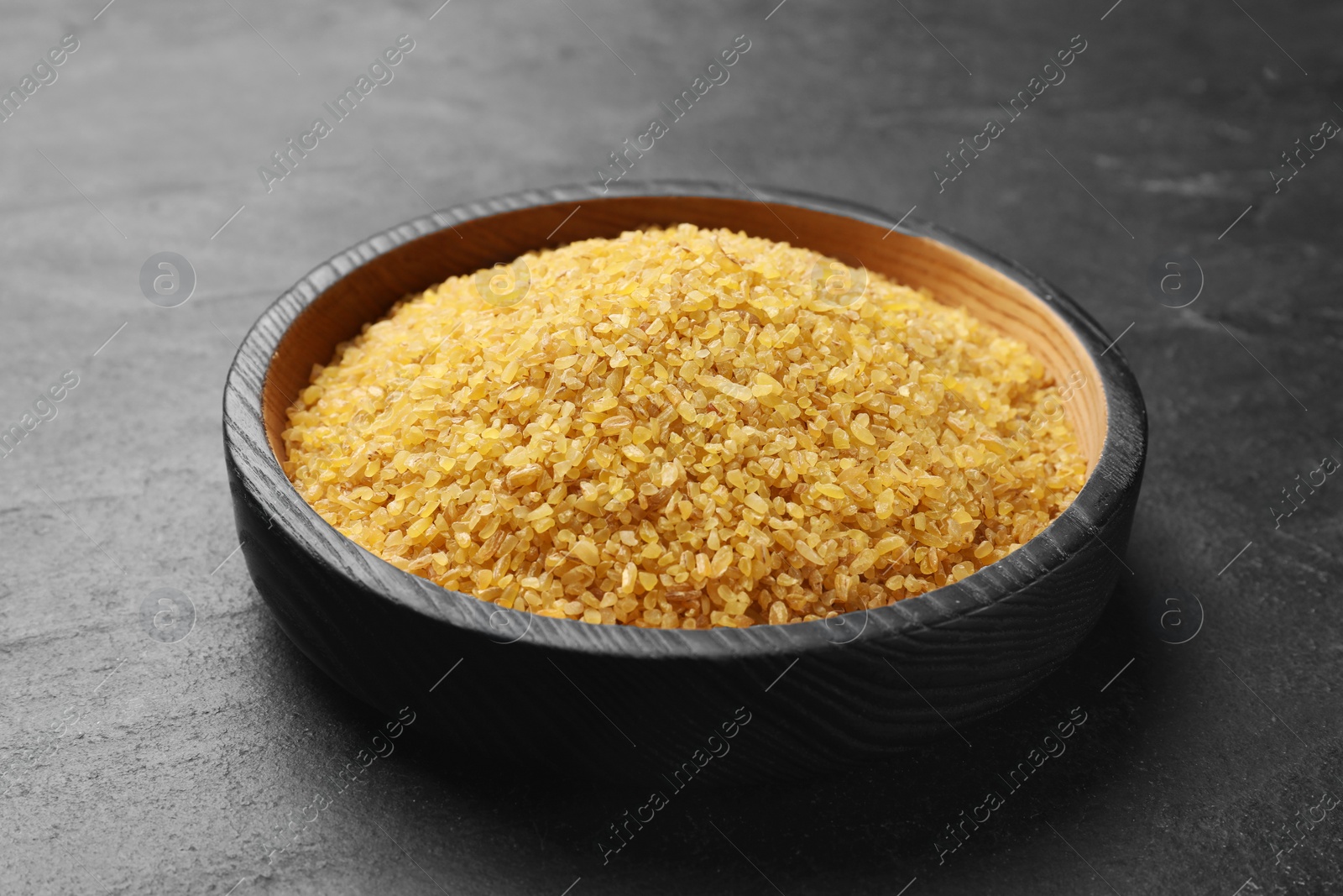 Photo of Raw bulgur in bowl on dark gray table, closeup