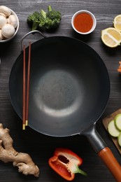 Photo of Empty iron wok and chopsticks surrounded by ingredients on dark grey wooden table, flat lay