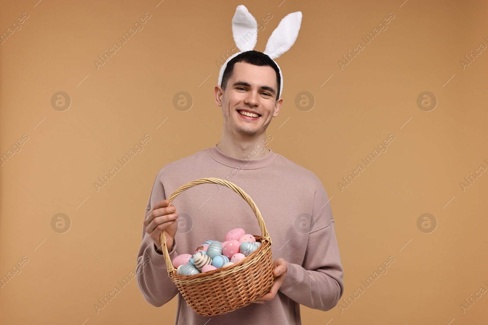 Photo of Easter celebration. Handsome young man with bunny ears holding basket of painted eggs on beige background