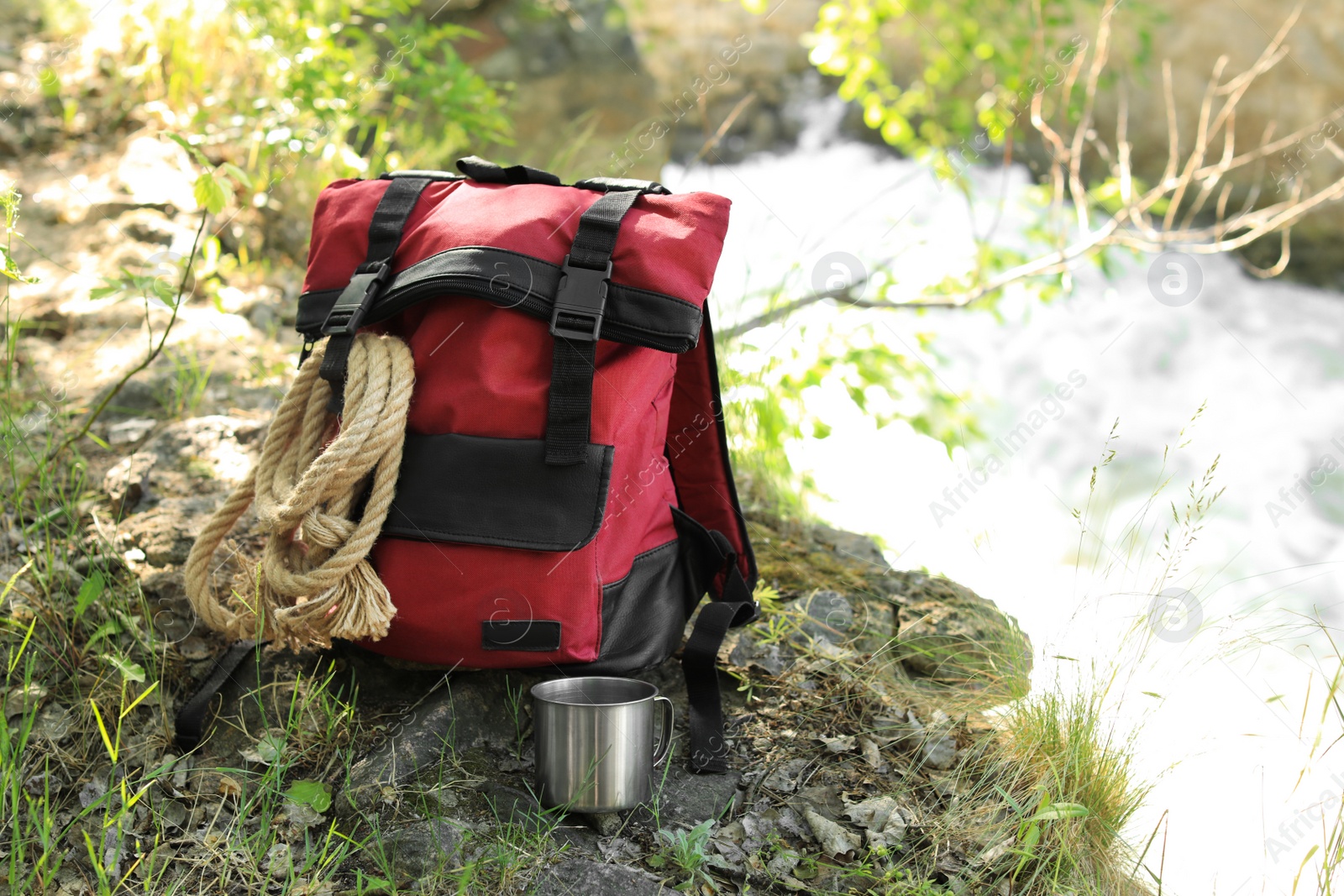 Photo of Camping backpack with rope and mug on stone near river. Space for text