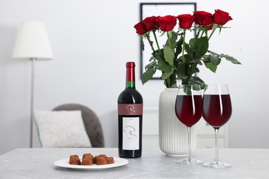 Photo of Bottle, glasses of red wine, chocolate truffles and vase with roses on table in room. Romantic date