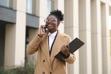 Photo of Happy woman with clipboard talking on smartphone outdoors. Lawyer, businesswoman, accountant or manager