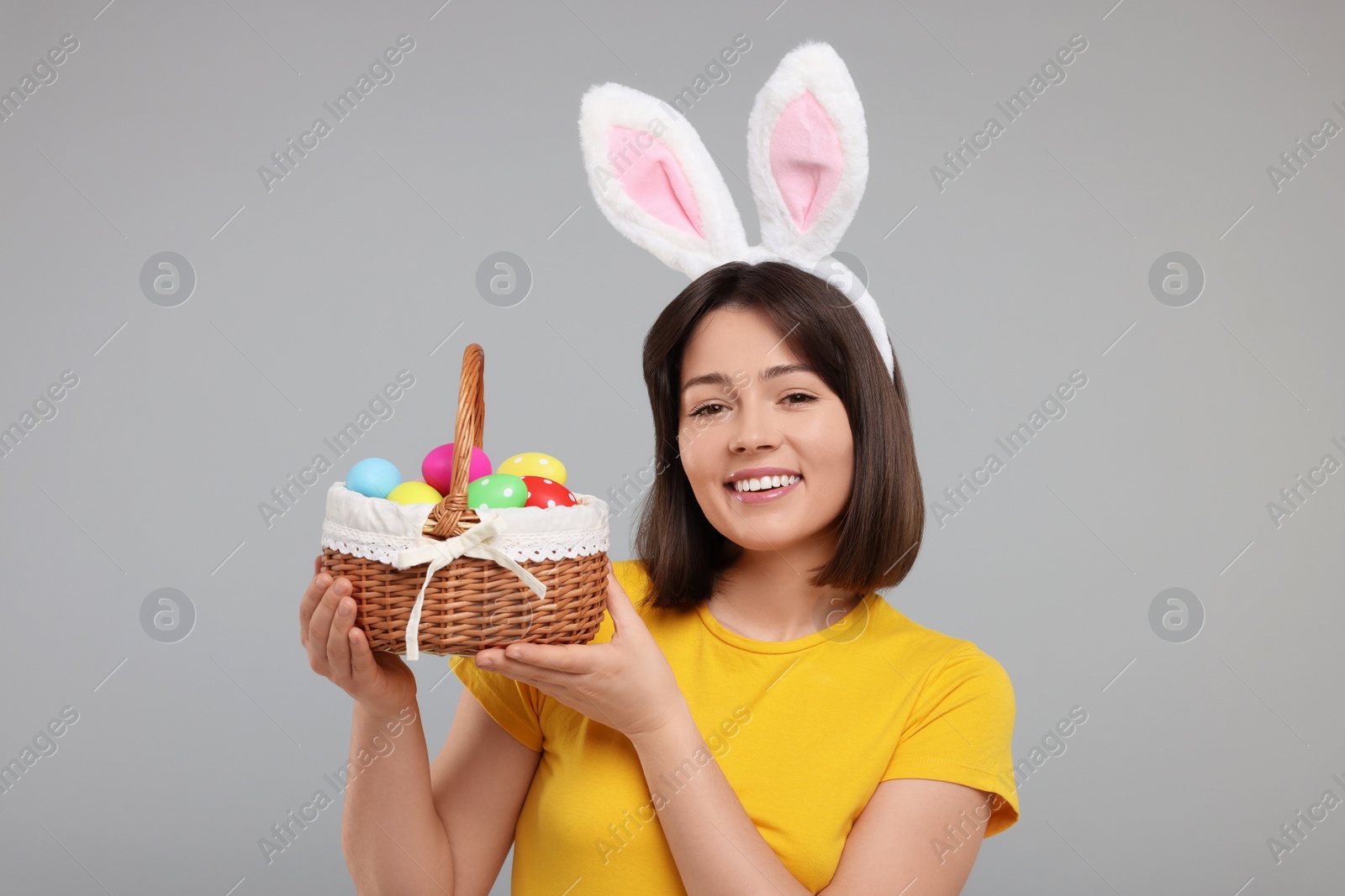 Photo of Easter celebration. Happy woman with bunny ears and wicker basket full of painted eggs on grey background