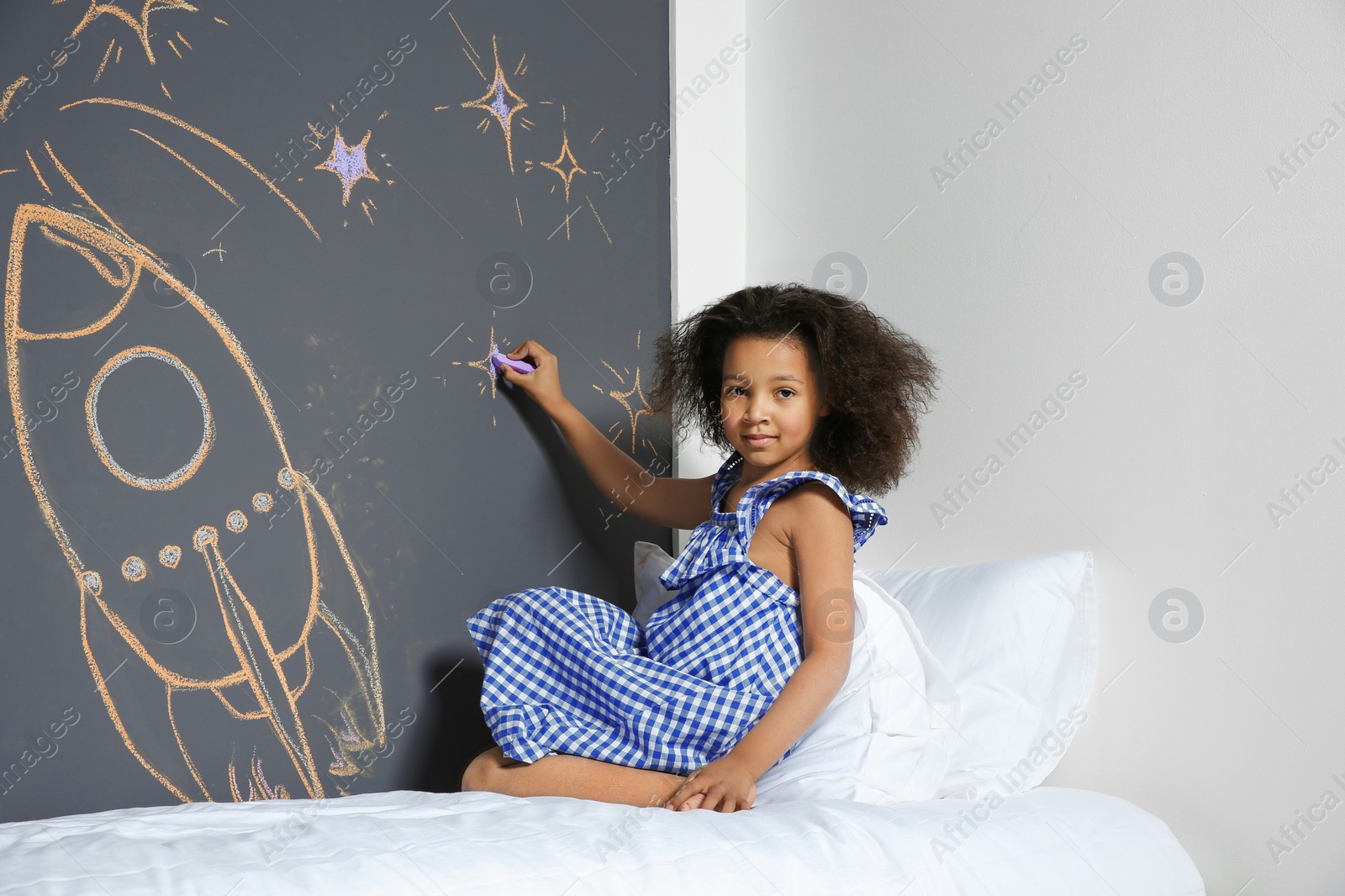 Photo of African-American child drawing rocket with chalk on wall in bedroom