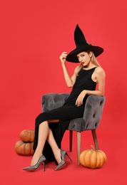 Beautiful woman wearing witch costume in armchair and pumpkins on red background. Halloween party