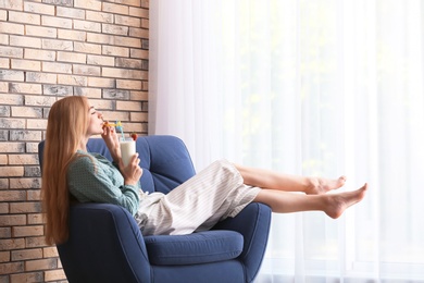 Young woman with glass of delicious milk shake in armchair indoors