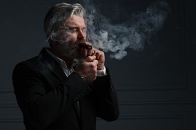 Bearded man lighting cigar on dark grey background. Space for text