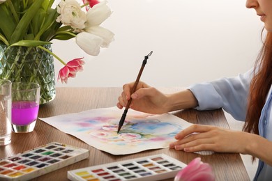 Photo of Woman painting flowers with watercolor at wooden table indoors, closeup. Creative artwork