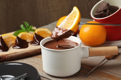 Dipping slice of orange into fondue pot with milk chocolate on wooden table