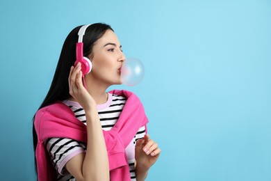 Photo of Fashionable young woman with headphones blowing bubblegum on light blue background, space for text