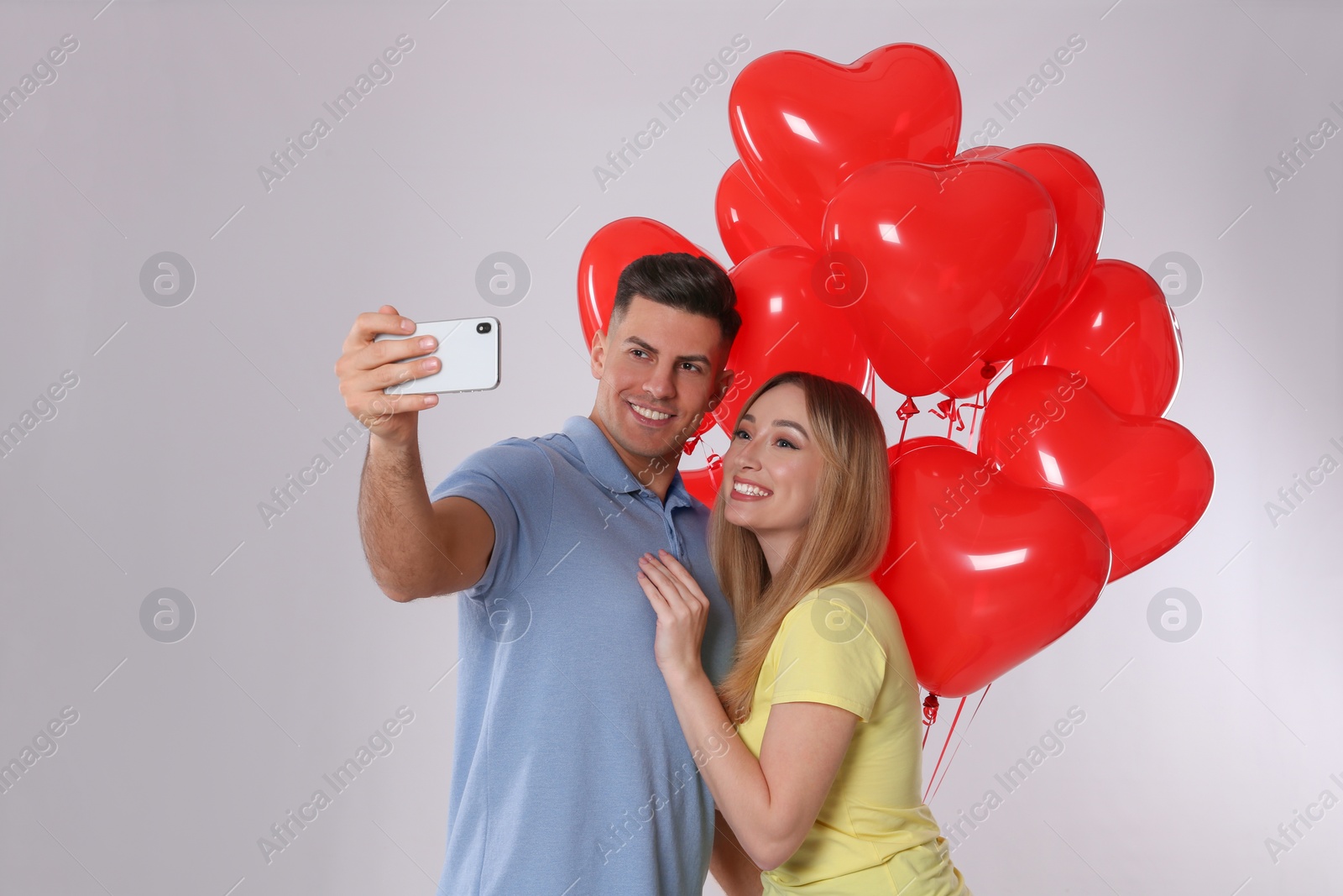 Photo of Lovely couple with heart shaped balloons taking selfie on light grey background. Valentine's day celebration