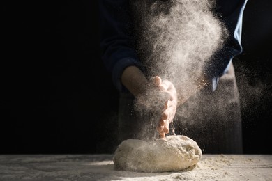 Photo of Making bread. Woman sprinkling flour over dough at table on dark background, closeup. Space for text