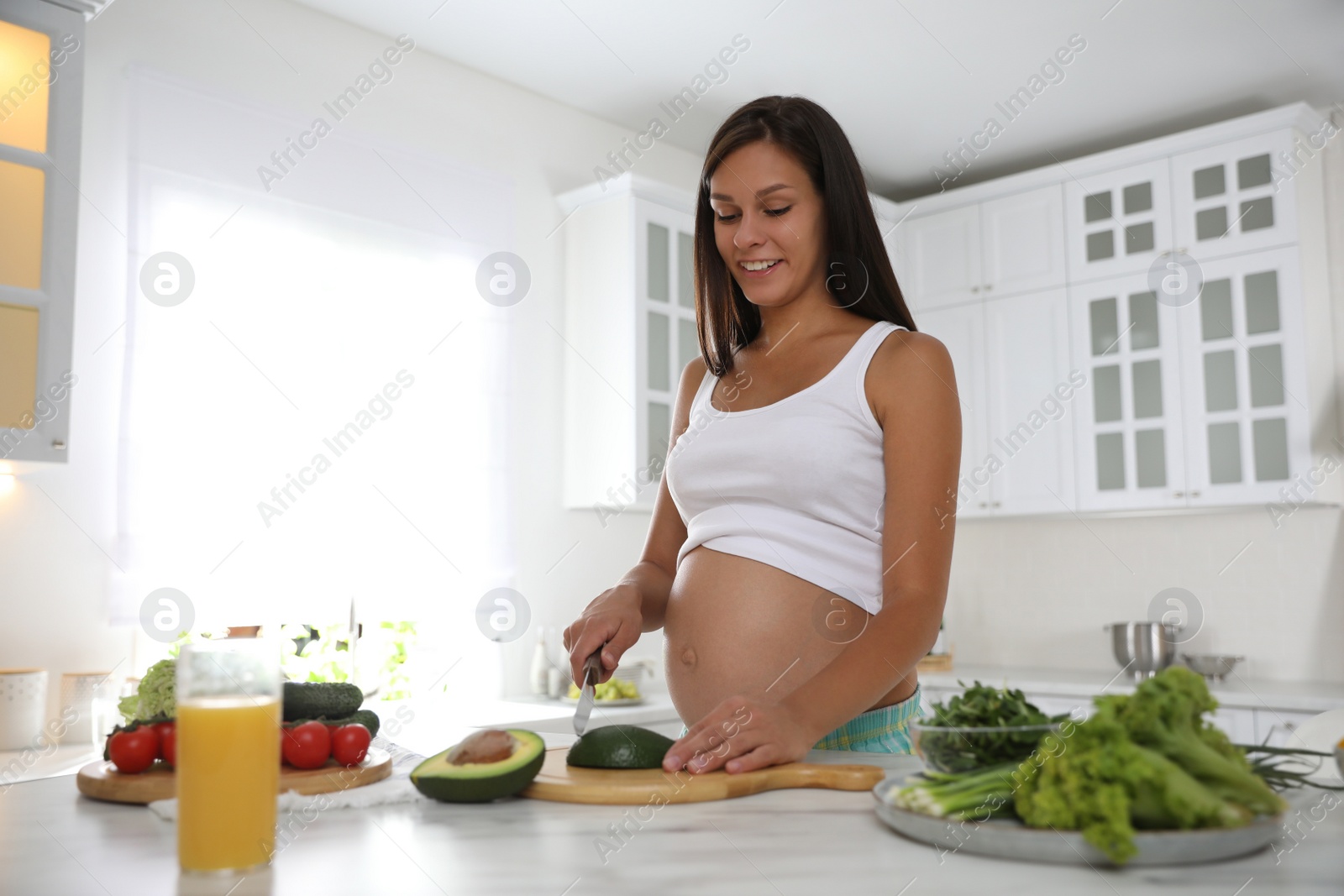 Photo of Young pregnant woman cutting avocado at table in kitchen. Taking care of baby health