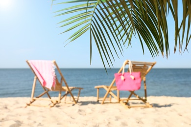 Photo of Blurred view of wooden sunbeds and beach accessories on sandy shore, focus on palm leaves