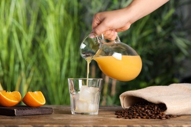 Photo of Woman pouring orange juice into glass with ice cubes at wooden table, closeup