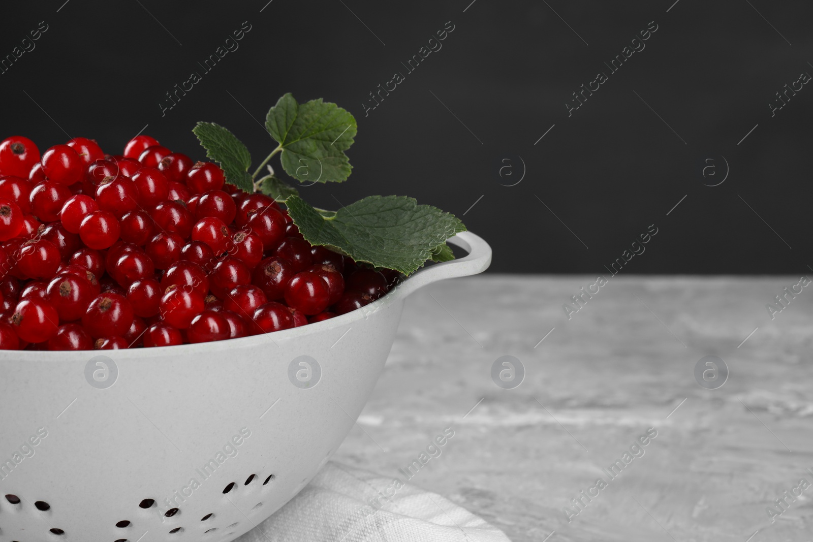 Photo of Ripe red currants and leaves in colander on grey textured table, closeup. Space for text