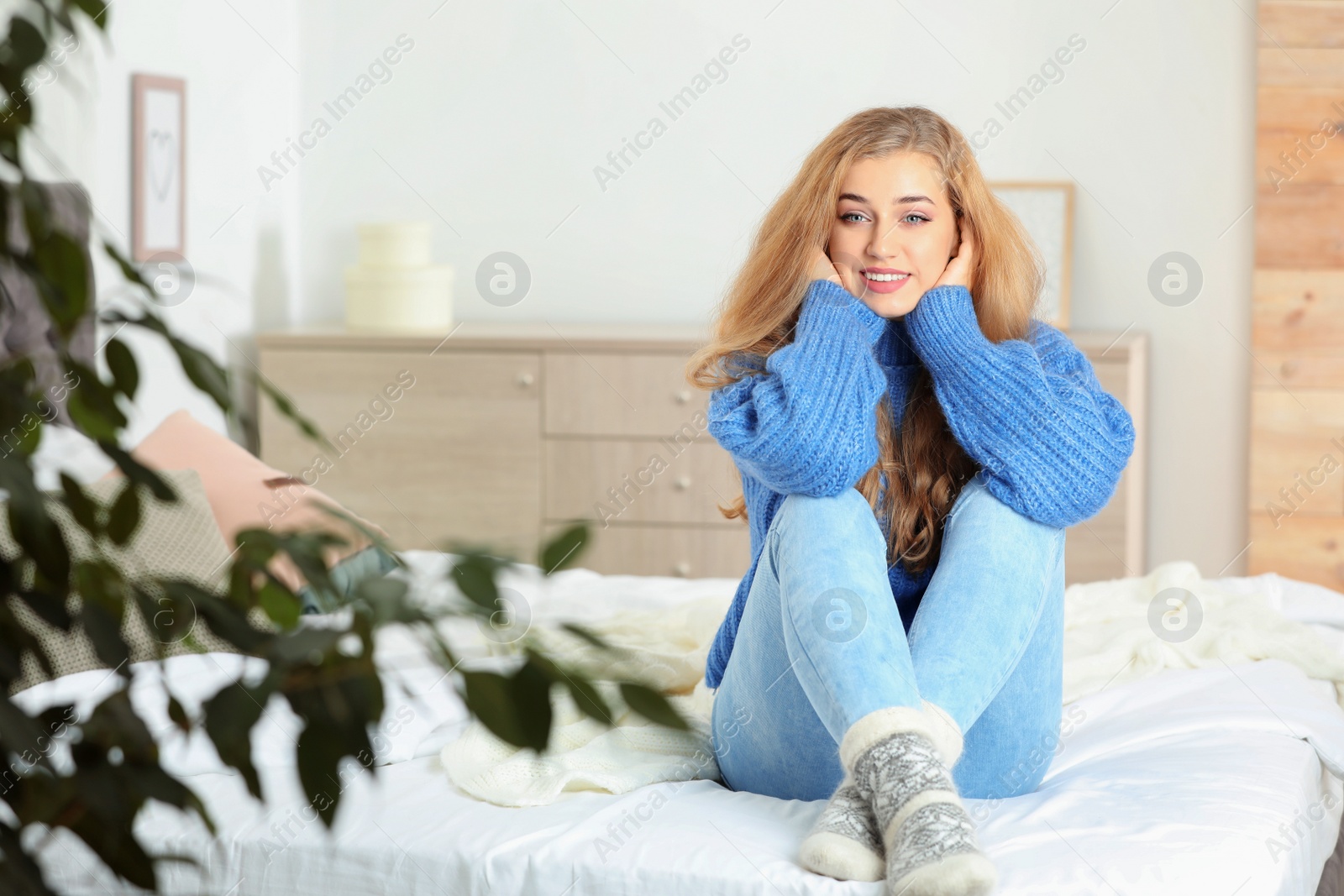 Photo of Beautiful smiling young woman in cozy warm sweater sitting on bed at home. Space for text