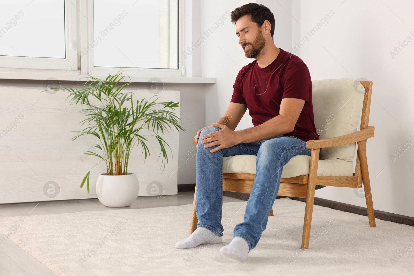 Photo of Man suffering from leg pain and touching knee on soft armchair at home