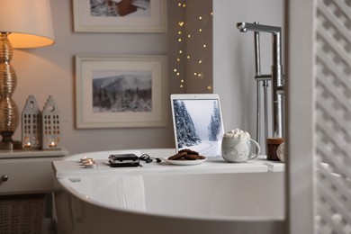 Photo of White wooden tray with tablet, cassette player and burning candles on bathtub in bathroom
