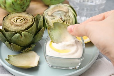 Photo of Woman dipping delicious cooked artichoke into sauce at table, closeup