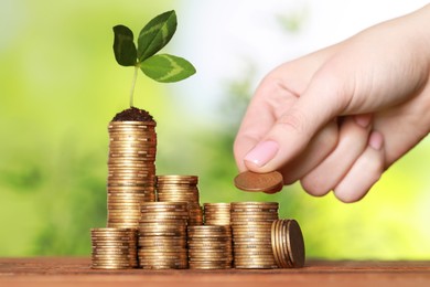 Woman putting coin onto stack with green sprout at wooden table against blurred background, closeup. Investment concept