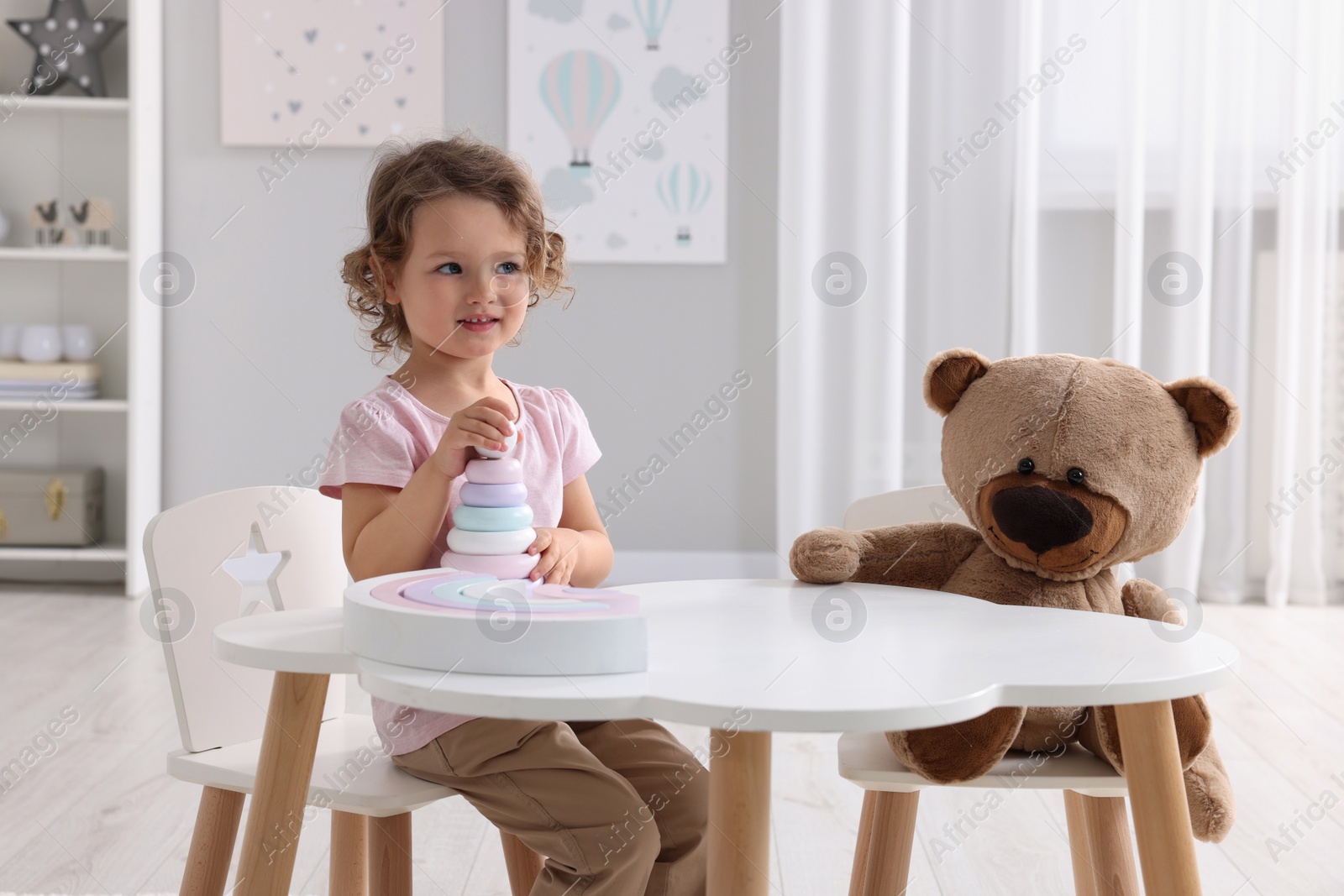 Photo of Cute little girl playing with toy and teddy bear at white table in room