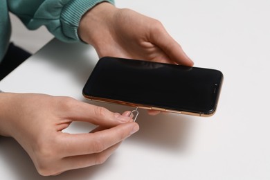 Woman with ejector opening SIM card tray in smartphone at white table, closeup