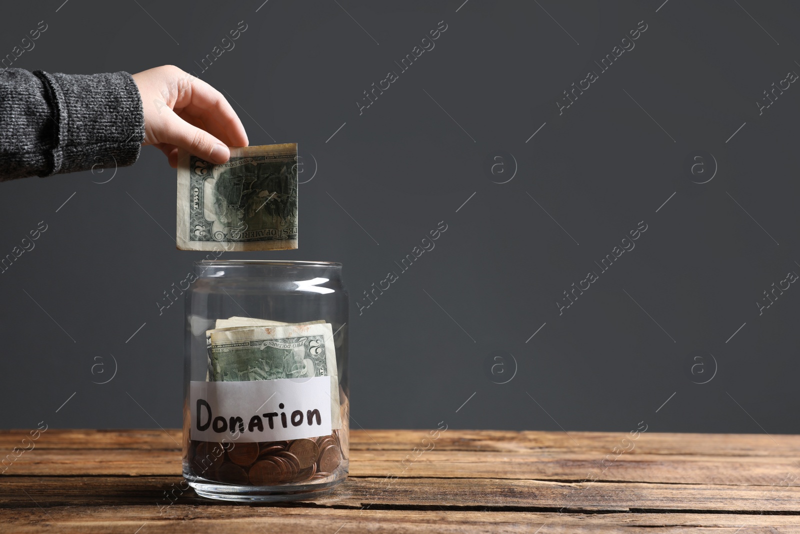 Photo of Woman putting money into donation jar on wooden table against grey background, closeup. Space for text