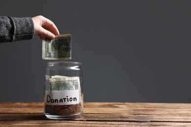 Photo of Woman putting money into donation jar on wooden table against grey background, closeup. Space for text