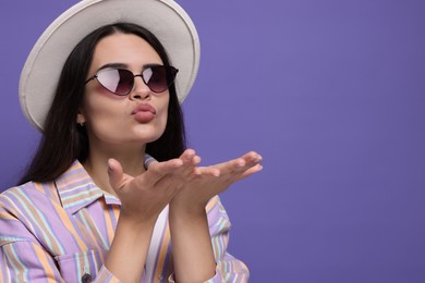 Photo of Beautiful young woman with stylish hat blowing kiss on purple background. Space for text