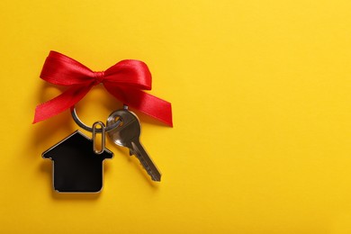 Photo of Key with trinket in shape of house and red bow on yellow background, top view. Space for text. Housewarming party