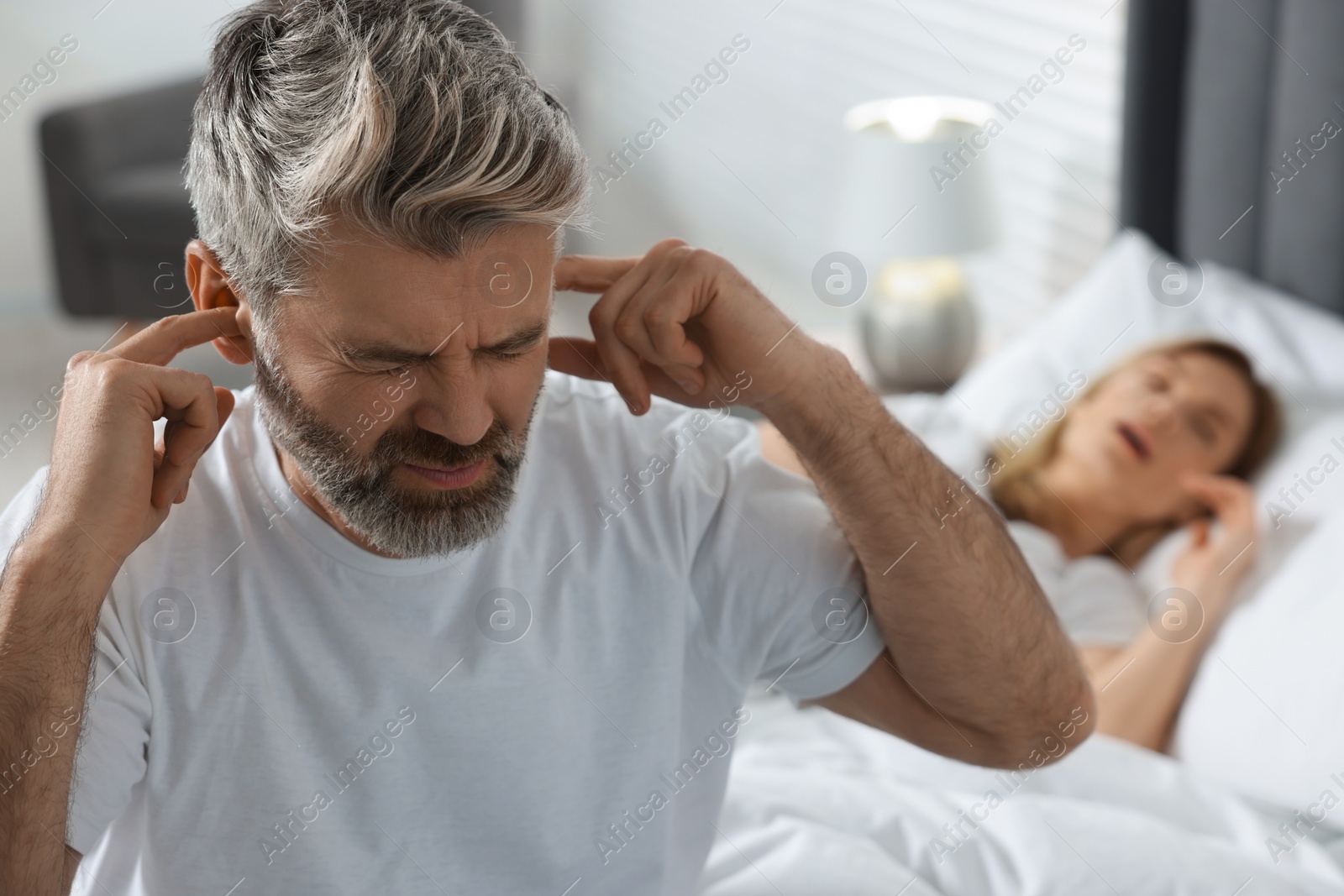 Photo of Irritated man covering his ears in bed at home, space for text. Problem with snoring wife