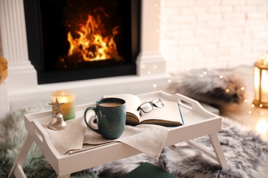 Photo of Cup of coffee, burning candle and book on tray near fireplace indoors. Cozy atmosphere