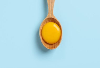 Wooden spoon with raw egg yolk on light blue background, top view