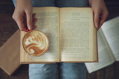 Woman with cup of coffee reading book indoors, top view