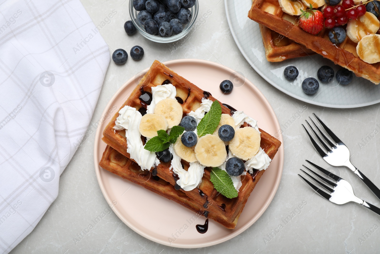 Photo of Plate of delicious Belgian waffles with blueberry, banana, whipped cream and chocolate sauce on light marble table, flat lay
