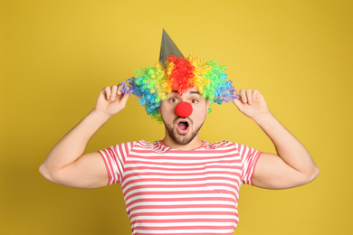 Photo of Emotional young man with party hat and clown wig on yellow background. April fool's day