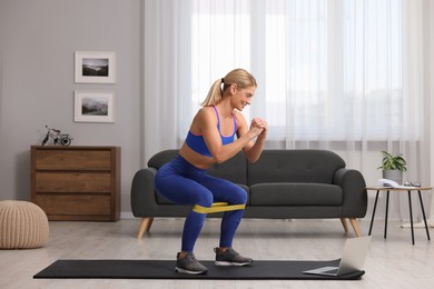 Photo of Fit woman doing squats with fitness elastic band near laptop on mat at home