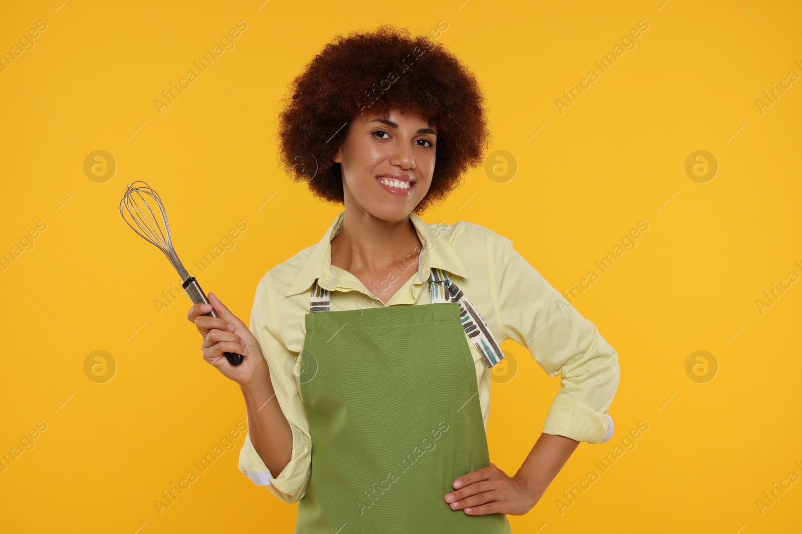 Photo of Happy young woman in apron holding whisk on orange background