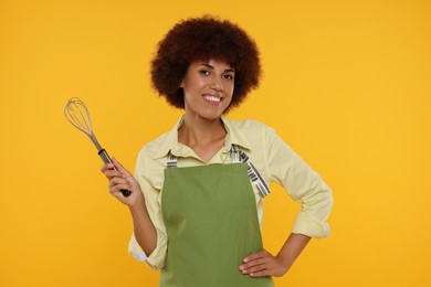 Happy young woman in apron holding whisk on orange background