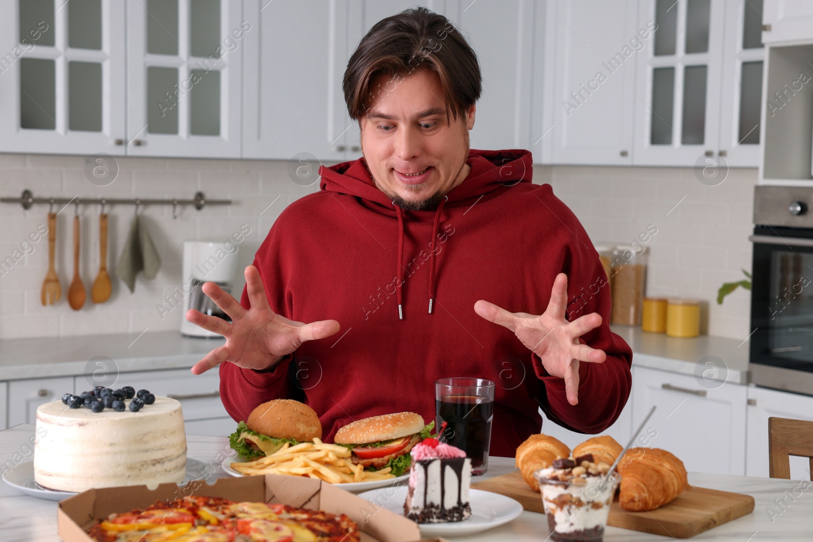 Photo of Hungry overweight man at table with sweets and fast food in kitchen