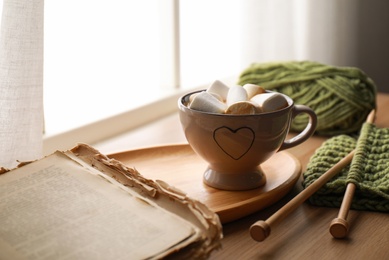 Cup of hot winter drink with marshmallows and knitting on wooden sill near window